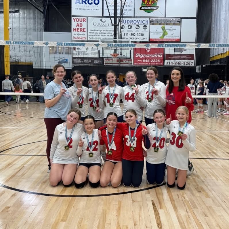 A 314 VBC team and two coaches kneeling and standing under the net at STL Crossfire, medals on and index fingers raised.