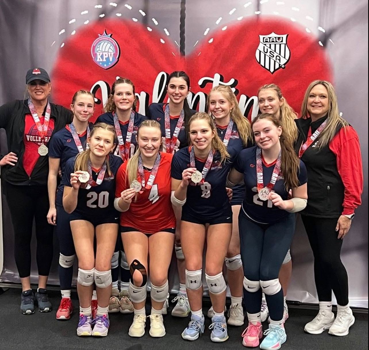 A 314 VBC team with two coaches holding their medals up to the camera in front of a red heart AAU Valentine's tournament backdrop.