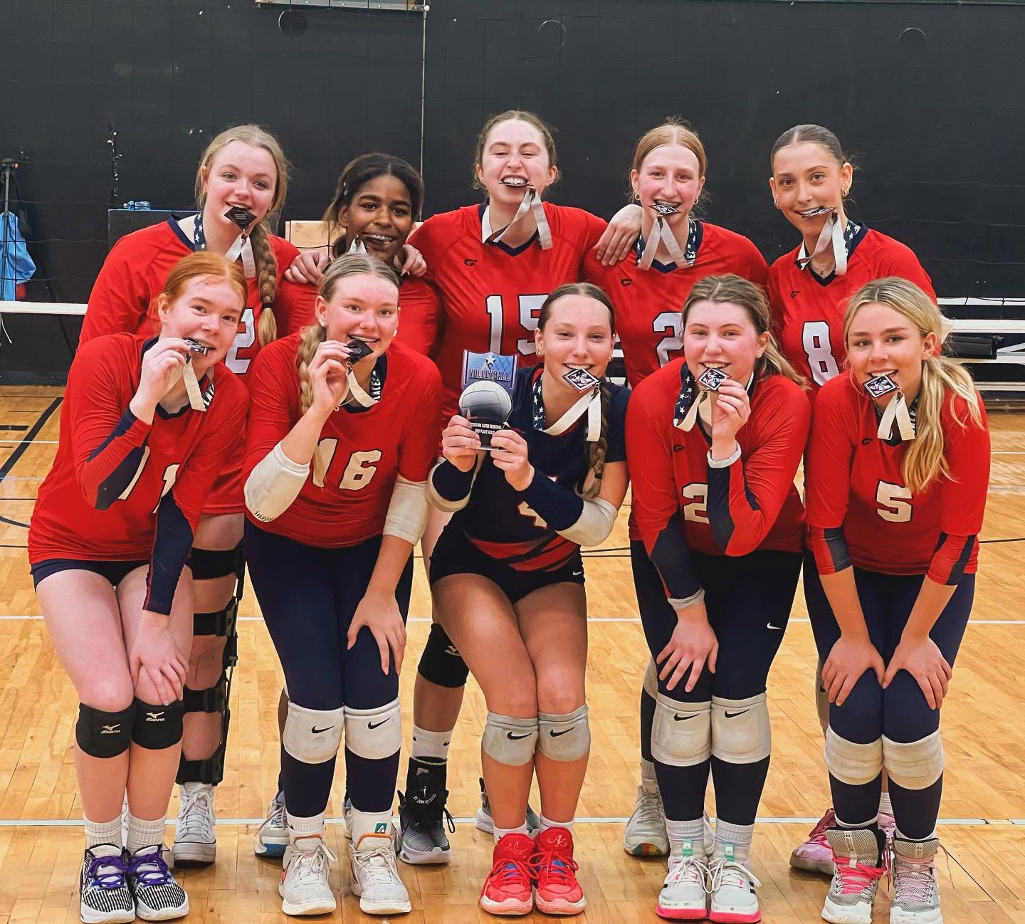 Ten 314 VBC players in red and navy jerseys crouched on the court, biting their medals, one holding a second-place trophy.