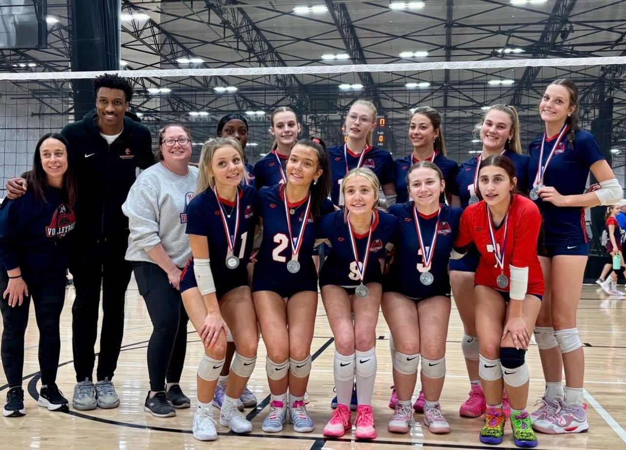 A 314 VBC team in navy and red jerseys with three coaches, all wearing silver medals, posed under the net on a convention centre court.