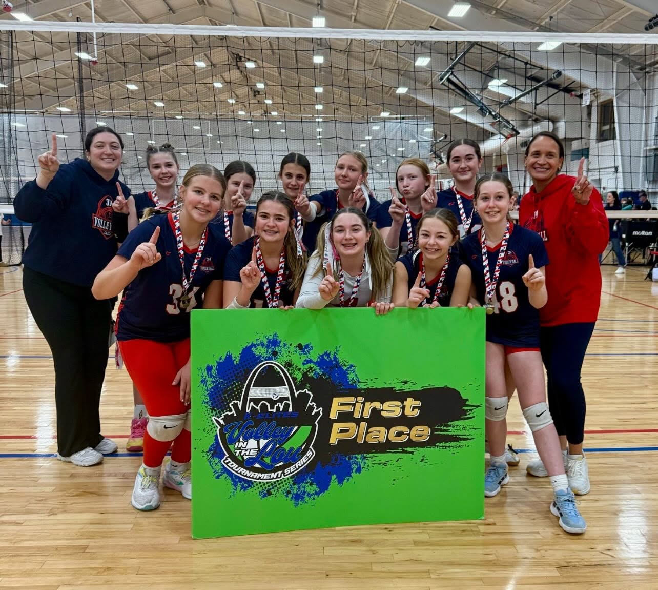 A 314 VBC team and two coaches on court behind a green First Place sign from the Volley in the Lou tournament series, everyone holding up one finger.